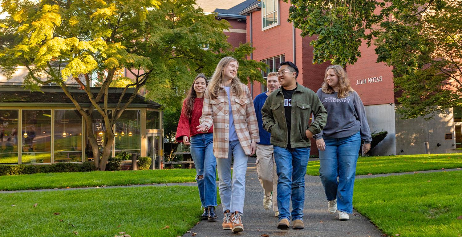 Five SPU students walking in SPU's Tiffany Loop, with the Student Union Building and Eaton Hall in the background.